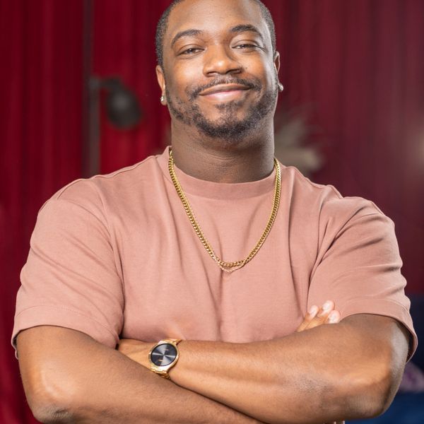 Man with arms crossed, wearing a gold chain and watch, smiles. In a room with red curtain.