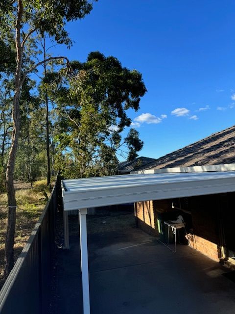 A carport with a metal roof next to a house with a dark roof and a fence. Trees and a blue sky are in the background. — Lee Pakau Roofing & Guttering in Tweed Heads, NSW