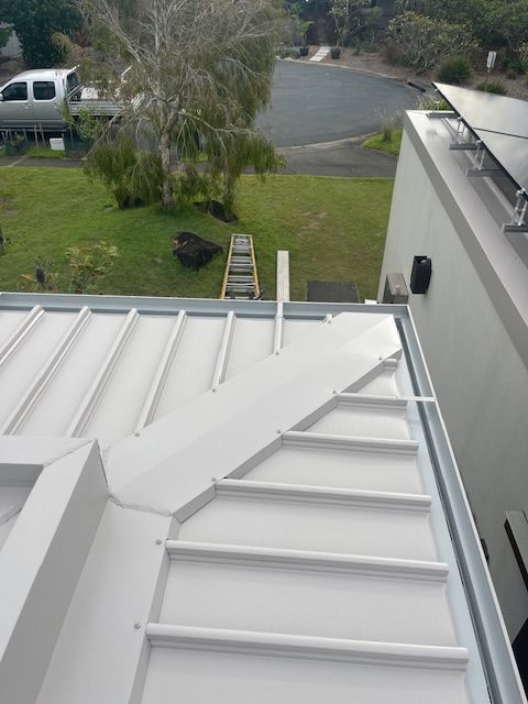 White metal roof with angled design, gutters, and partial solar panels. Ladder and street visible in the background. — Lee Pakau Roofing & Guttering in Banora Point, NSW