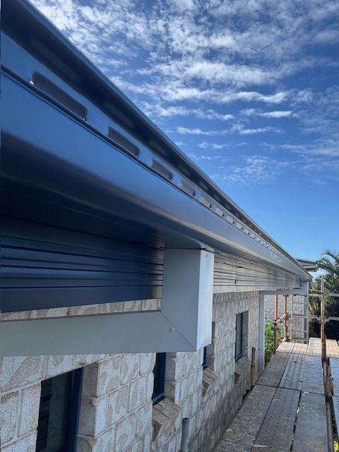 Dark Gray Gutters Along a Brick Building With a Blue Sky Background — Lee Pakau Roofing & Guttering in Banora Point, NSW