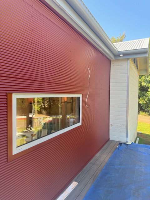 Red corrugated metal building with a rectangular window, white trim, and a small deck. — Lee Pakau Roofing & Guttering in Banora Point, NSW