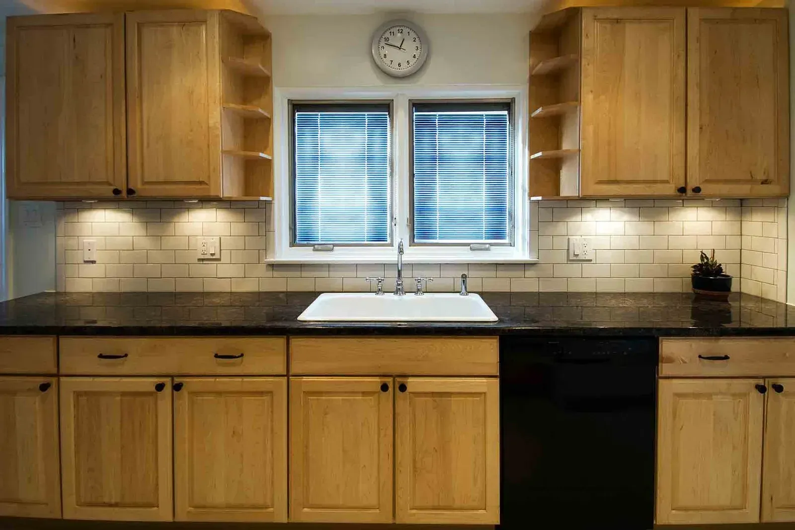 Kitchen with light wood cabinets, black countertop, and white sink under a window.