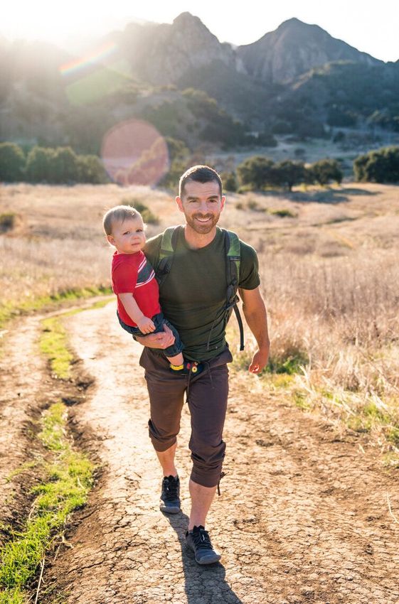 A man is carrying a baby in a carrier while walking down a dirt path.