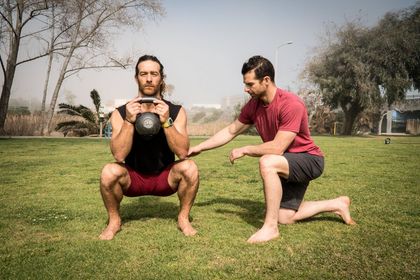 Two men are squatting in the grass with a kettlebell.