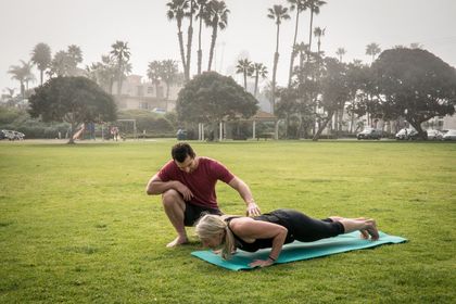 A man is helping a woman do push ups on a yoga mat in a park.