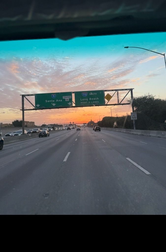 Highway at sunset, cars driving, sky with orange and pink hues, overhead street signs.