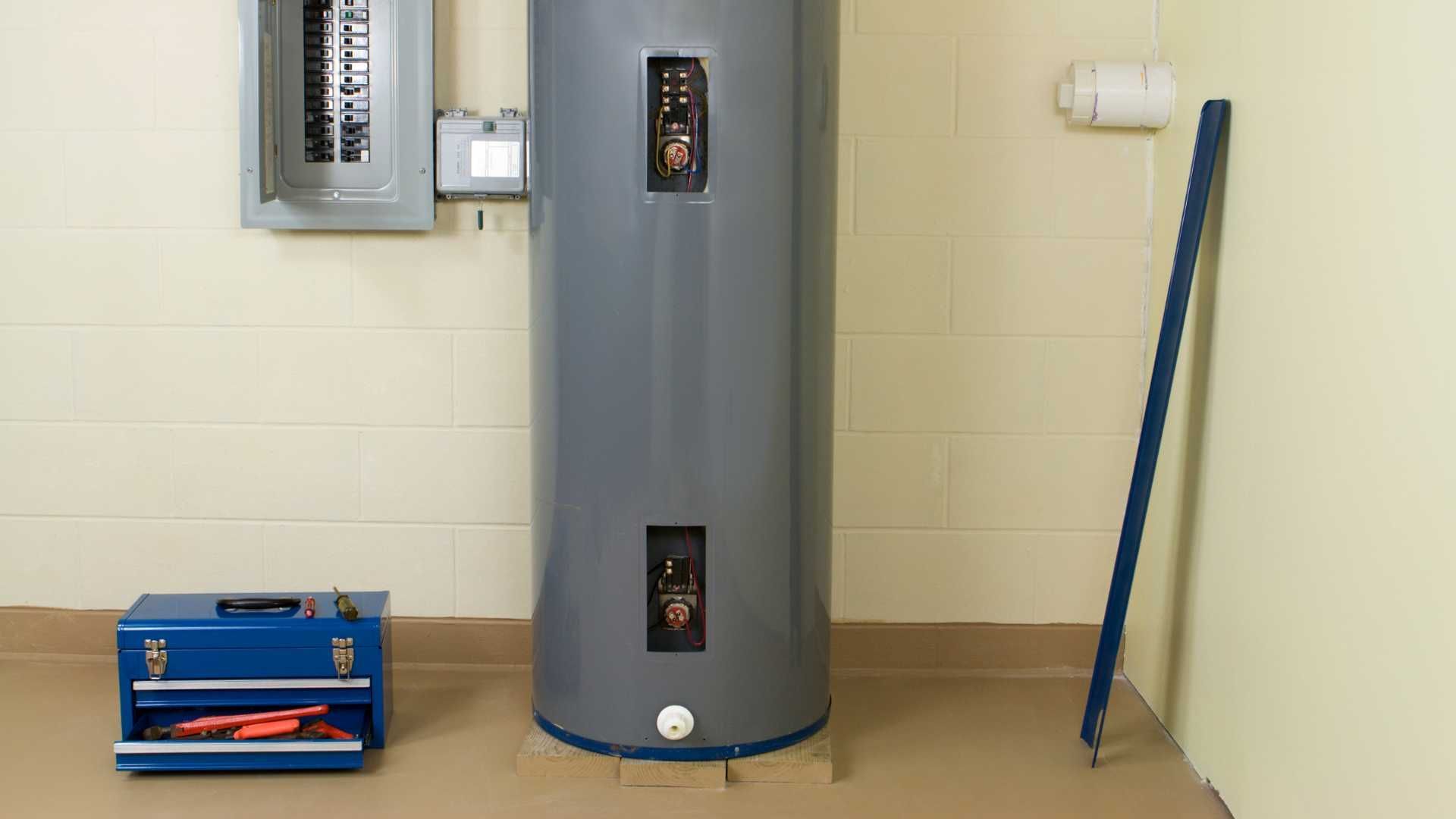 A water heater is sitting in a basement next to a toolbox.