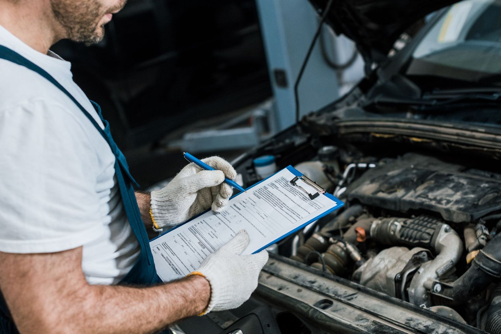 A man is writing on a clipboard while looking under the hood of a car.
