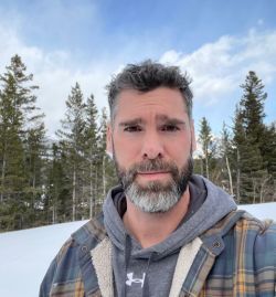 A man with a beard and a plaid jacket is standing in the snow.