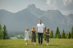 A man and three girls are standing in a grassy field with mountains in the background.