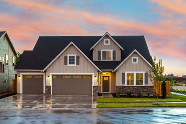Suburban house at sunset with lit garage and wet driveway, reflected in the rain-soaked street