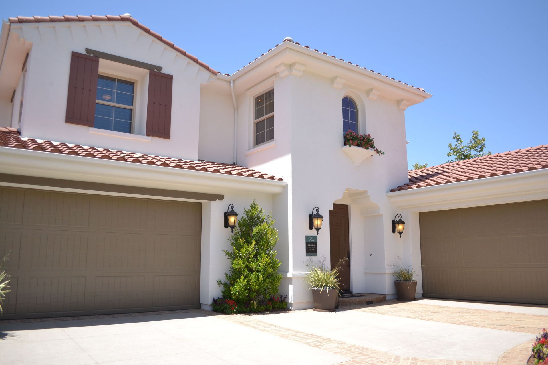 Two-story Mediterranean-style house with beige garage doors, white stucco, and red tile roof under a blue sky