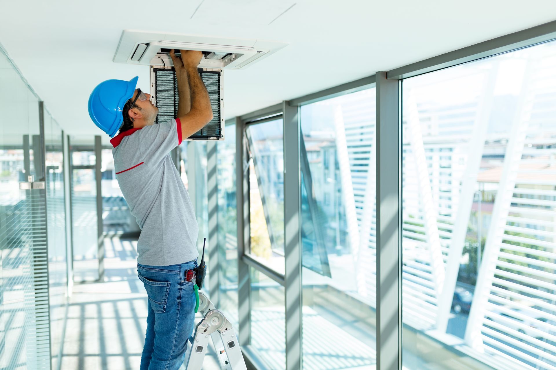 A technician in a blue hard hat repairs an air conditioning unit in a bright office space.