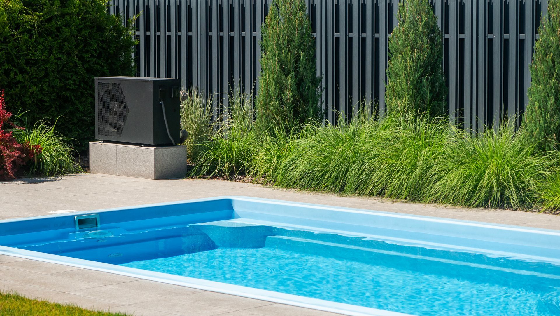 Poolside view: a black heat pump atop a stone base near a blue pool. A green hedge and fence are in the background.