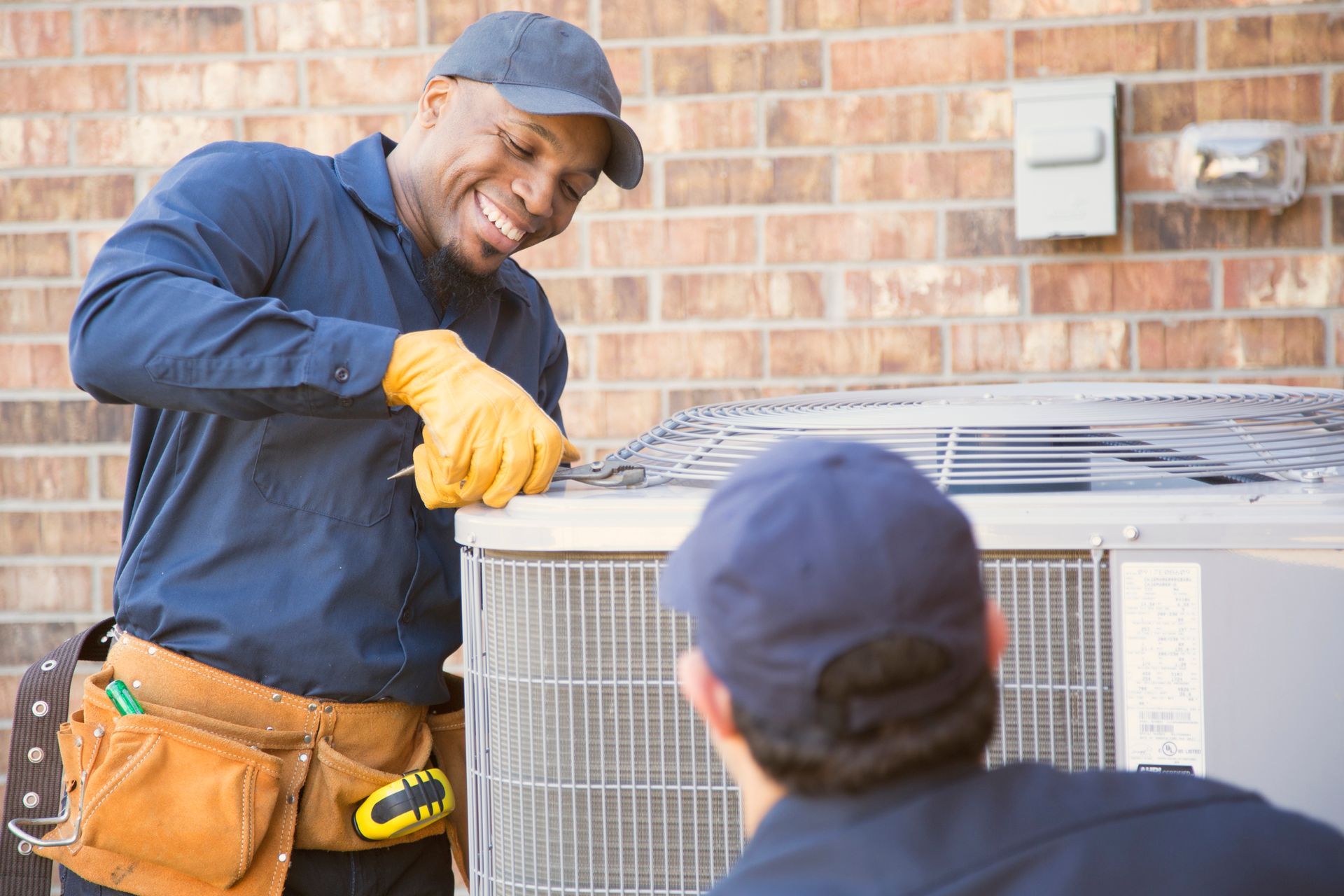 Two HVAC technicians working on an air conditioning unit outdoors. One smiles, wearing gloves and using a tool.