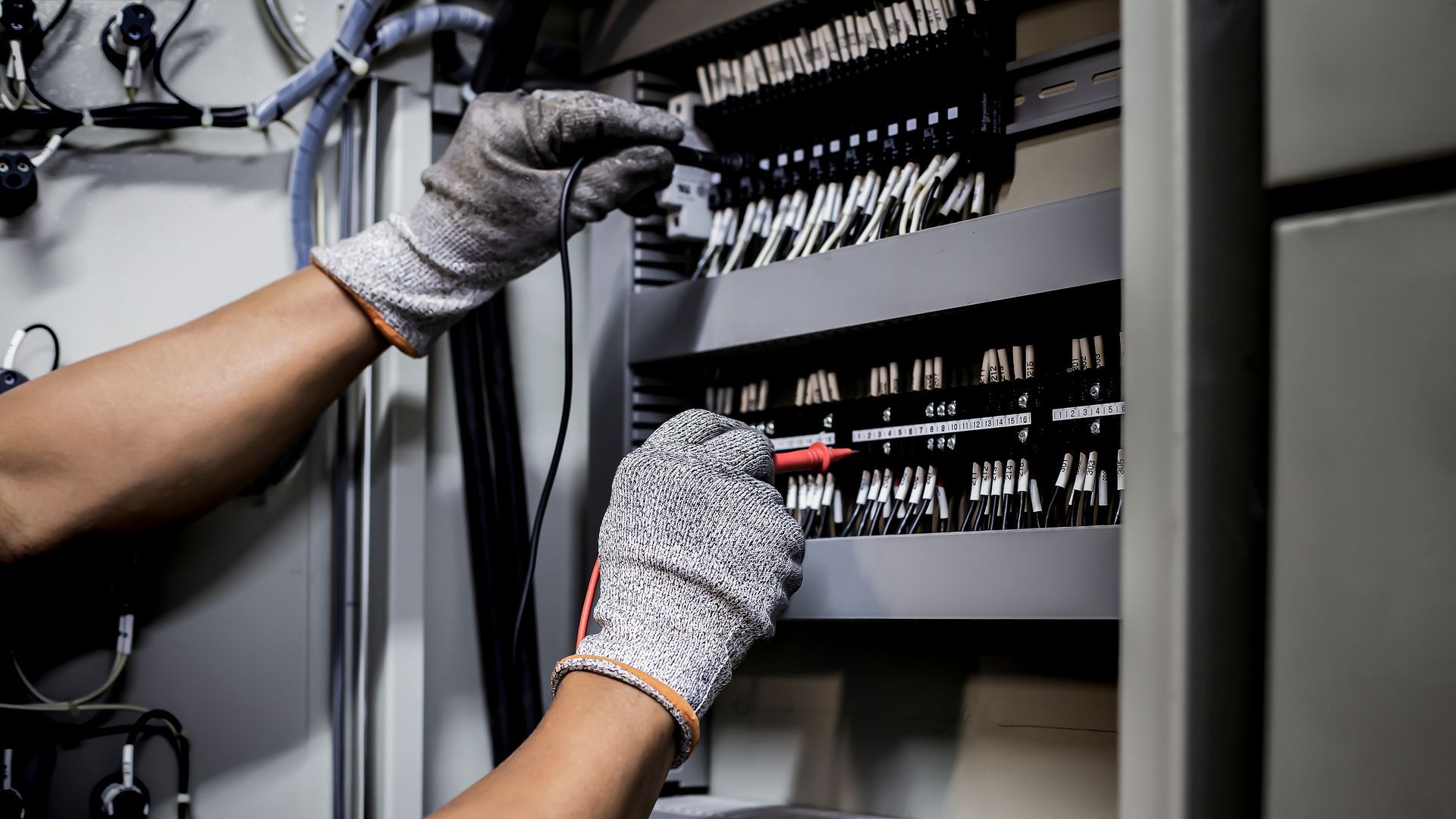 A person wearing blue gloves uses a multimeter to test a circuit breaker panel.