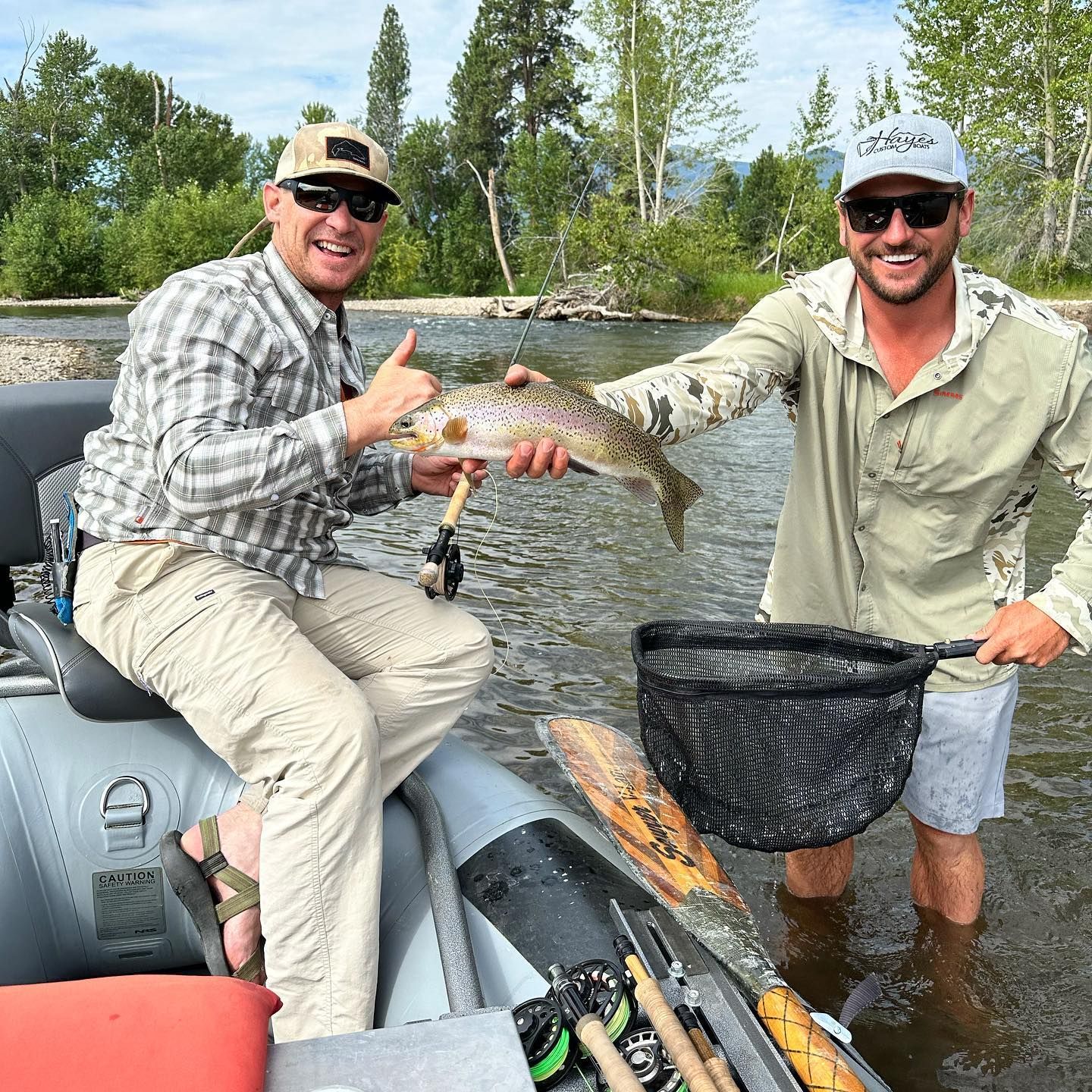 Two men fishing, holding a trout and a net in a river, one giving a thumbs up.