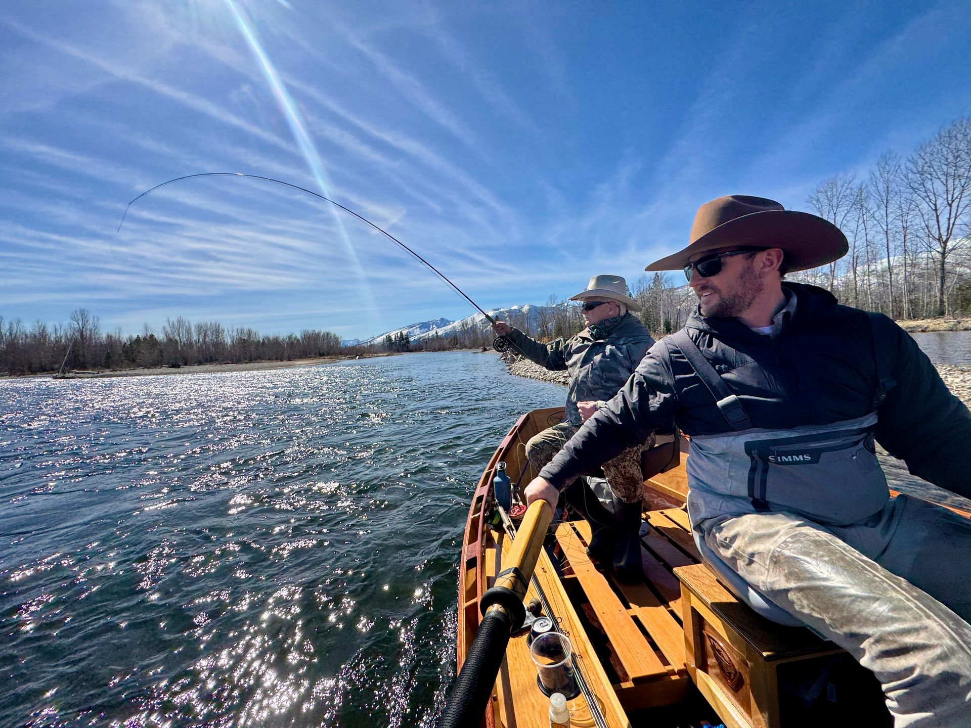 Two people fly fishing from a wooden boat on a river, sunny day.