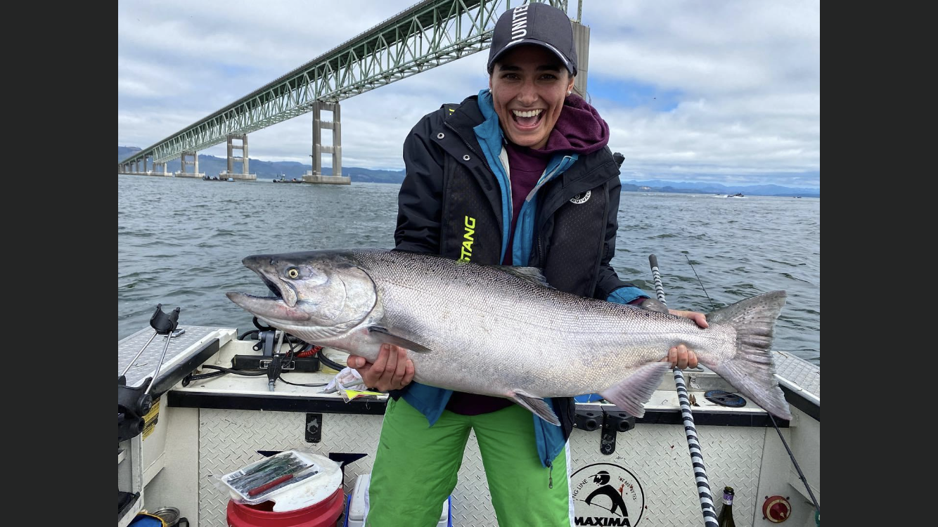 Woman on a boat holding a large, silver fish, smiling broadly, with a bridge in the background.