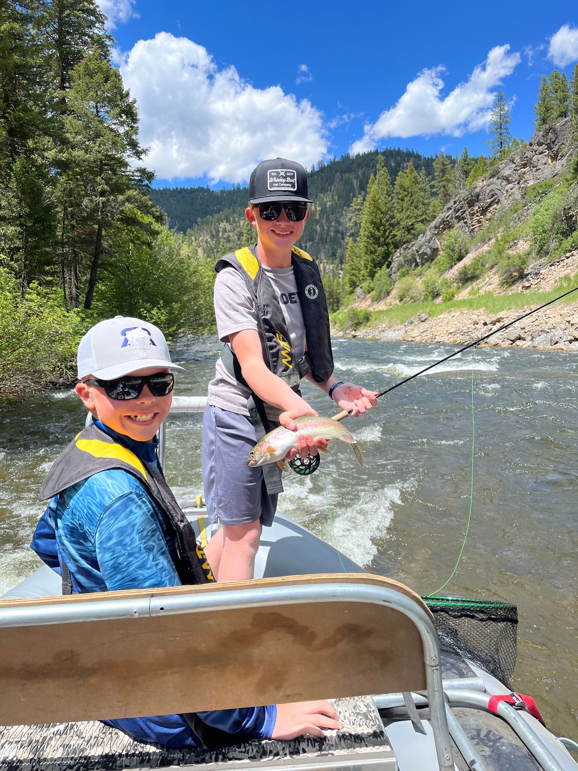 Two boys fishing from a boat on a river, one holding a fish. Sunny day, green trees, mountains.