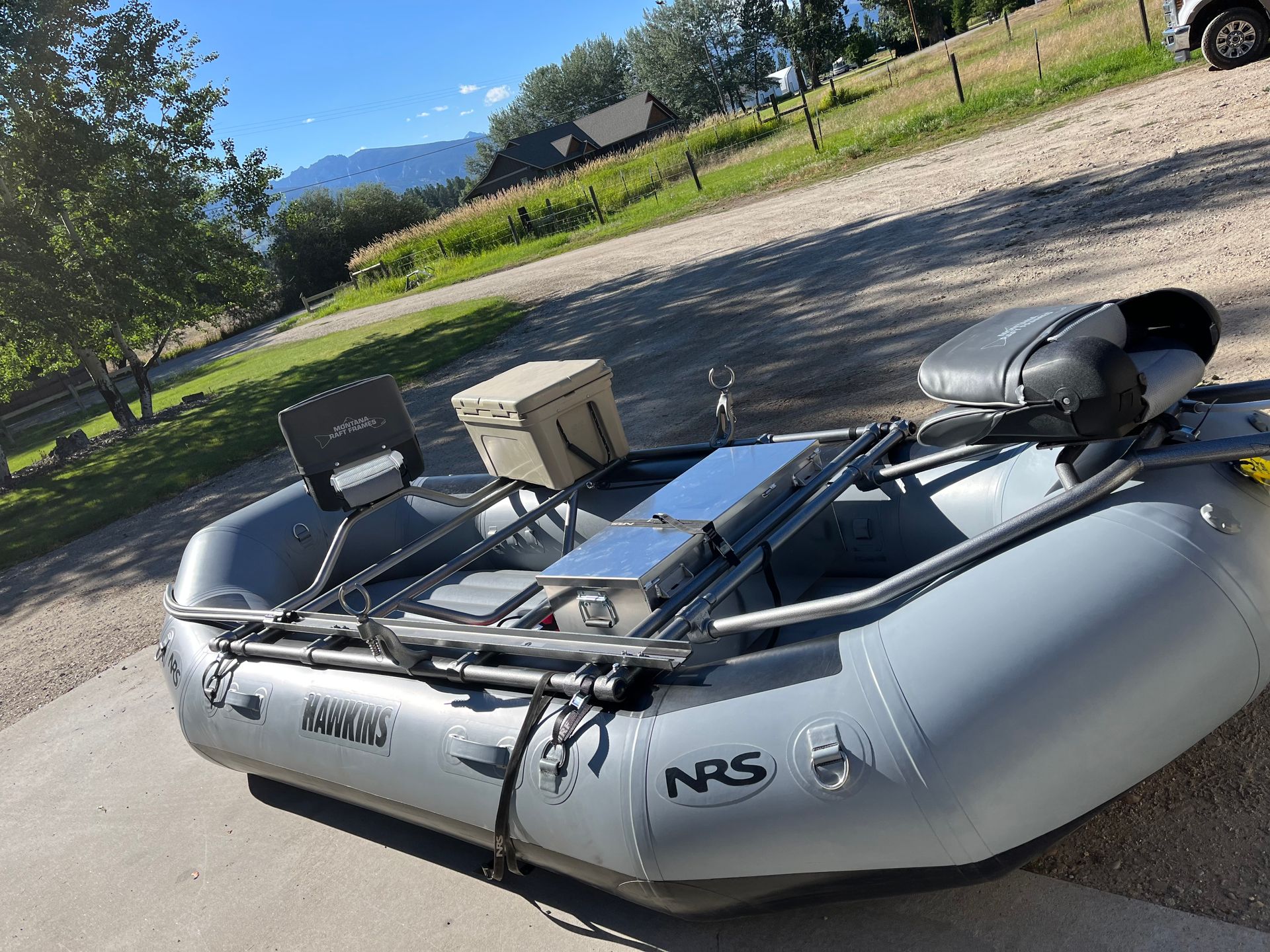 Gray NRS raft on a driveway, equipped with seats, cooler, and gear, mountains in background.