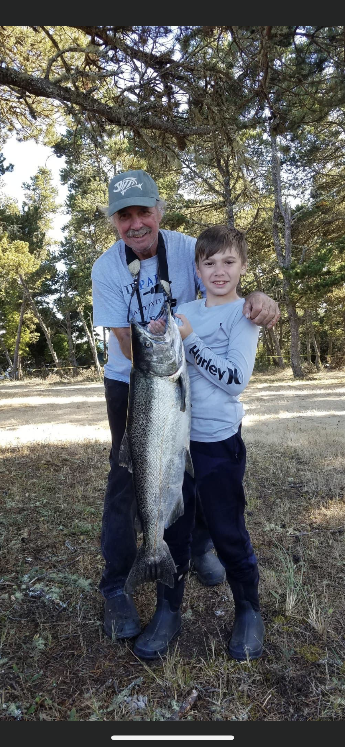 A man and boy smiling, holding a large fish, standing outside near trees and brown ground.