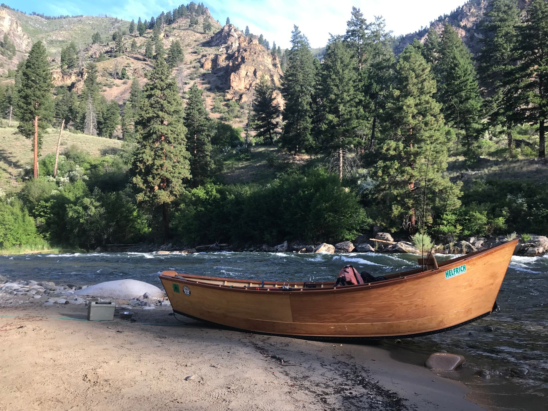 Wooden drift boat on a sandy riverbank, mountains and trees in the background.