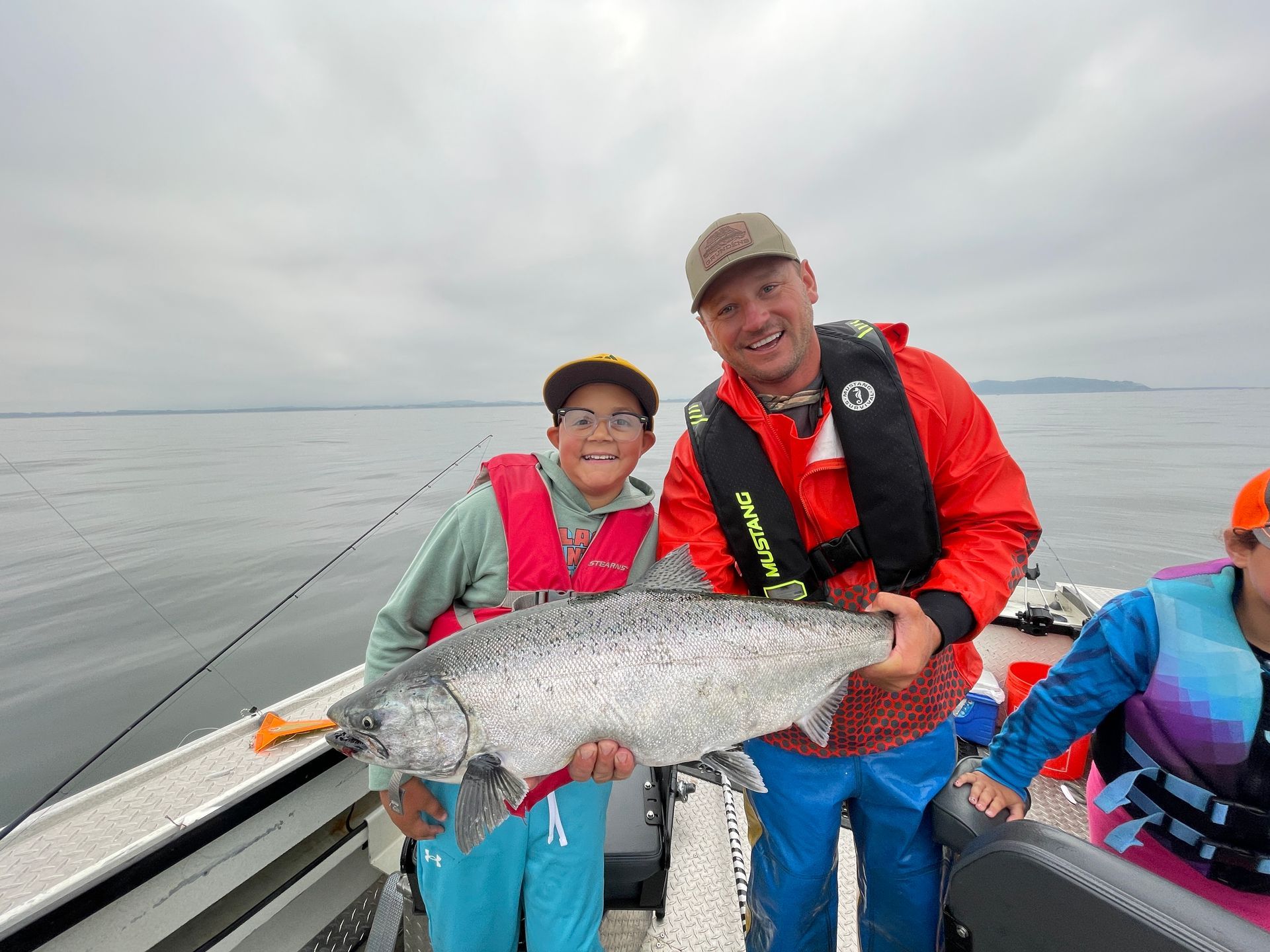 Boy and man on boat hold a large silver fish; both wearing life vests, smiling. Gray sky, water.