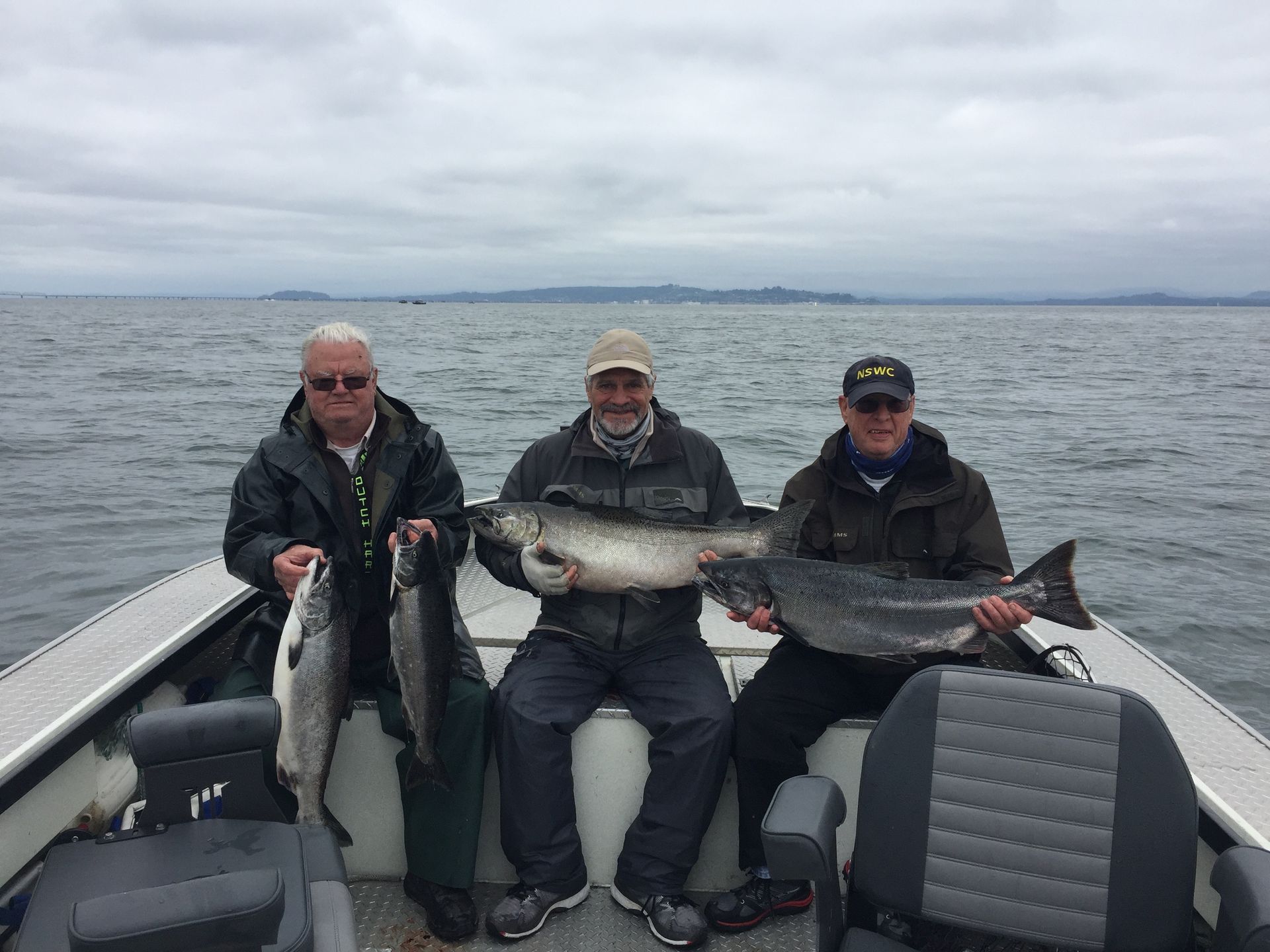 Three people on a boat holding large fish they caught, against an overcast sky over the water.