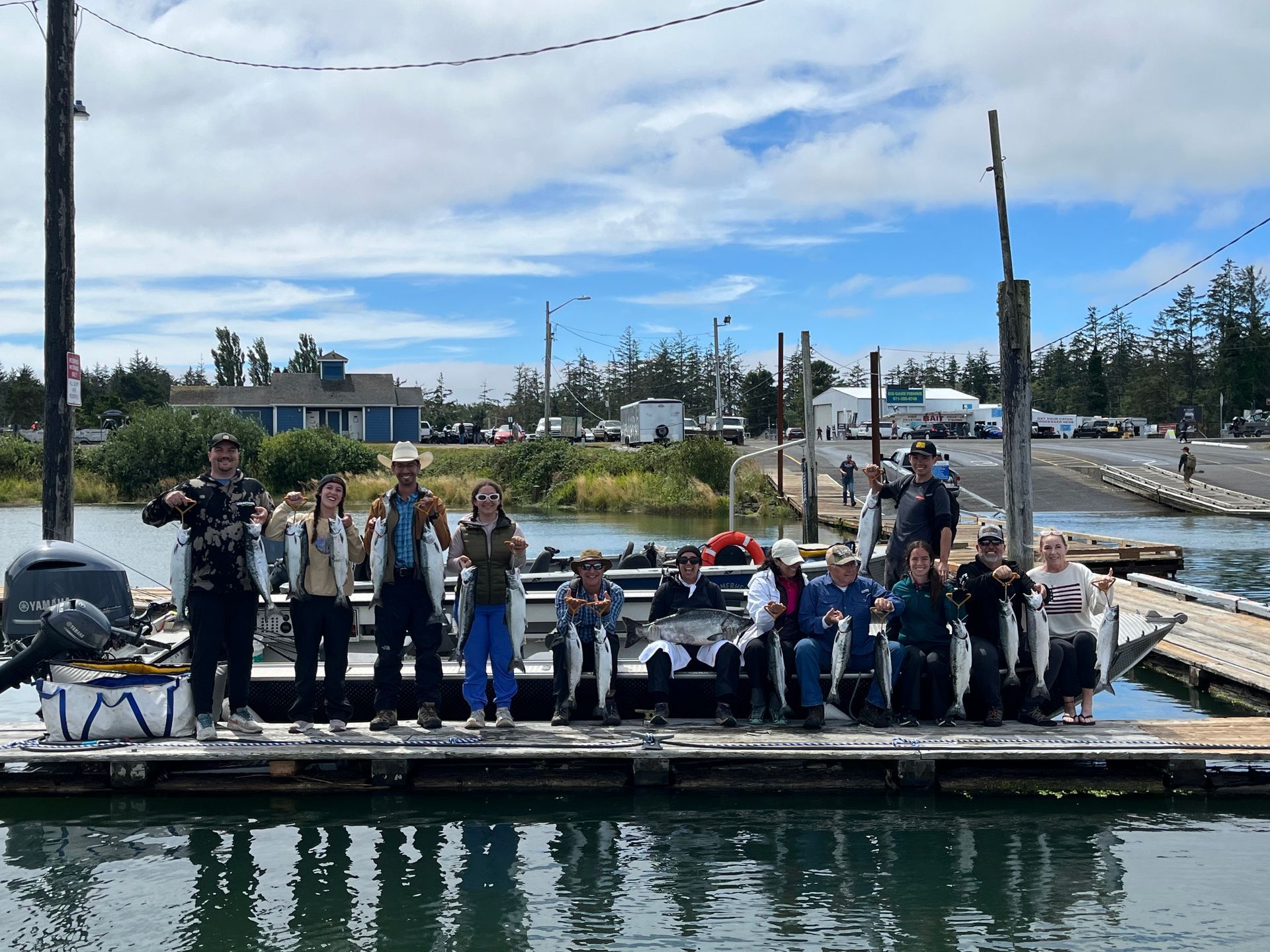 People on a dock hold up fish they caught, blue sky, water, and buildings in the background.