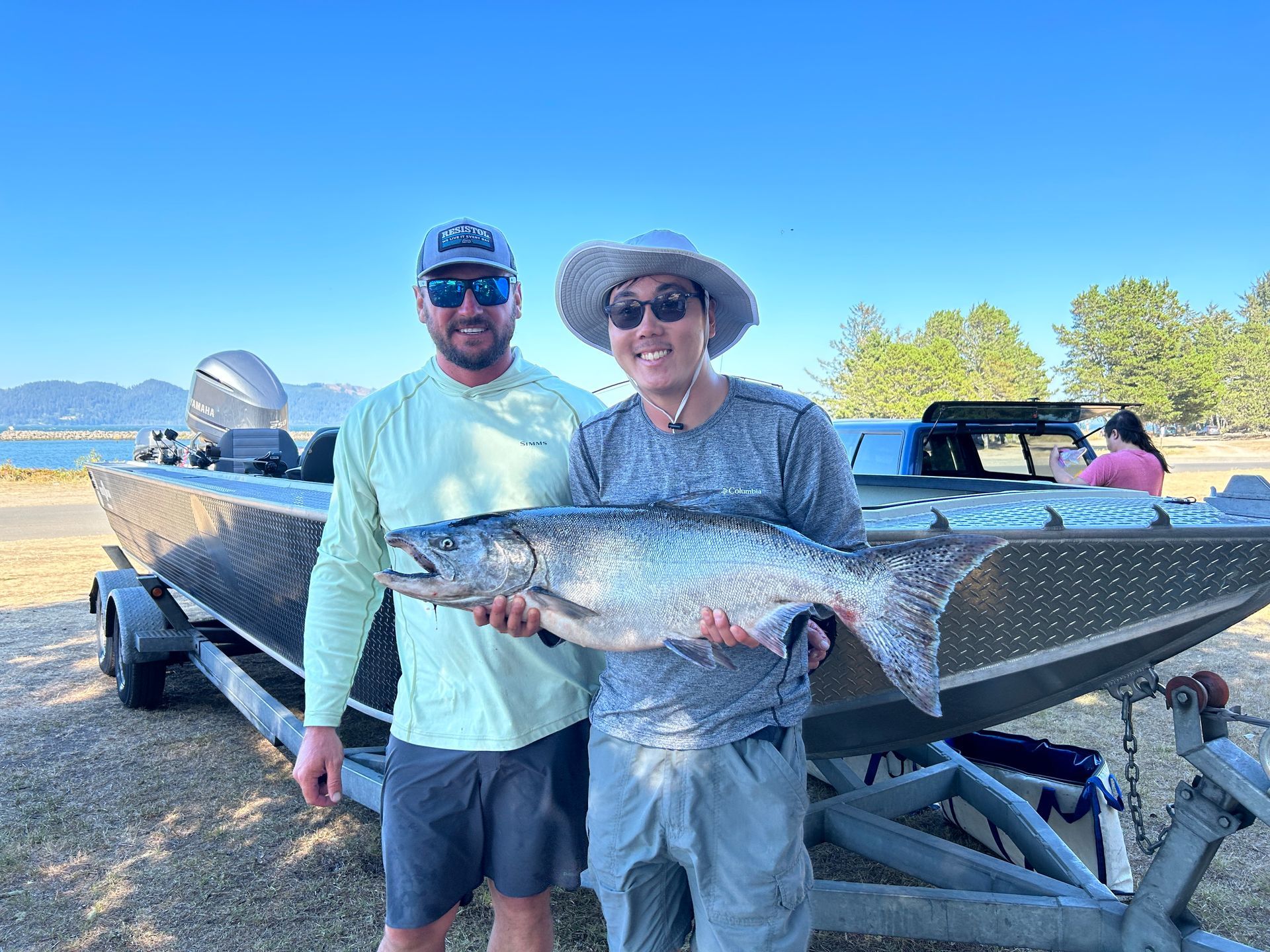 Two people pose with a large fish in front of a boat near a body of water.