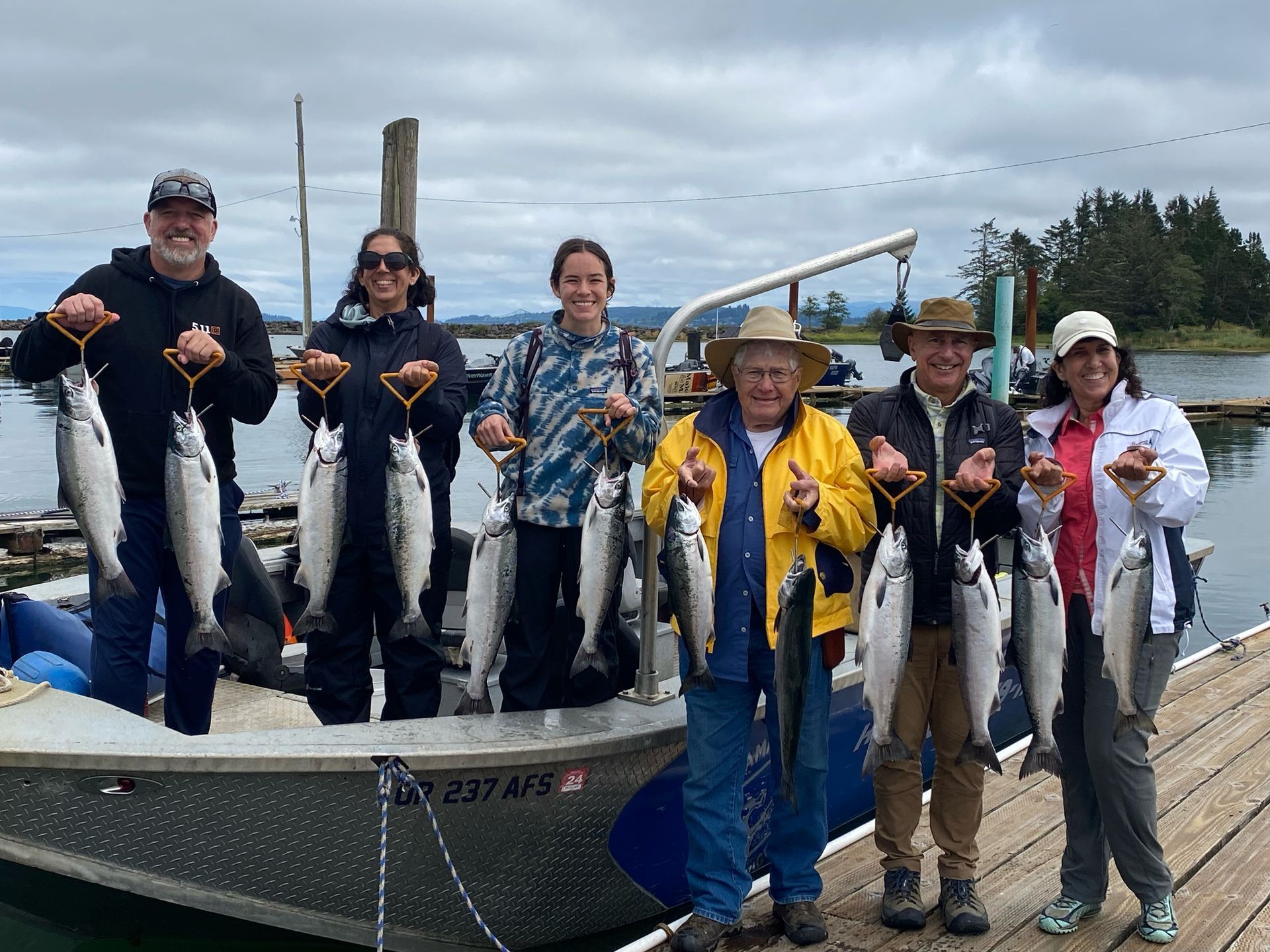 Group of people on a boat holding up several silver fish they caught, near a dock and water.