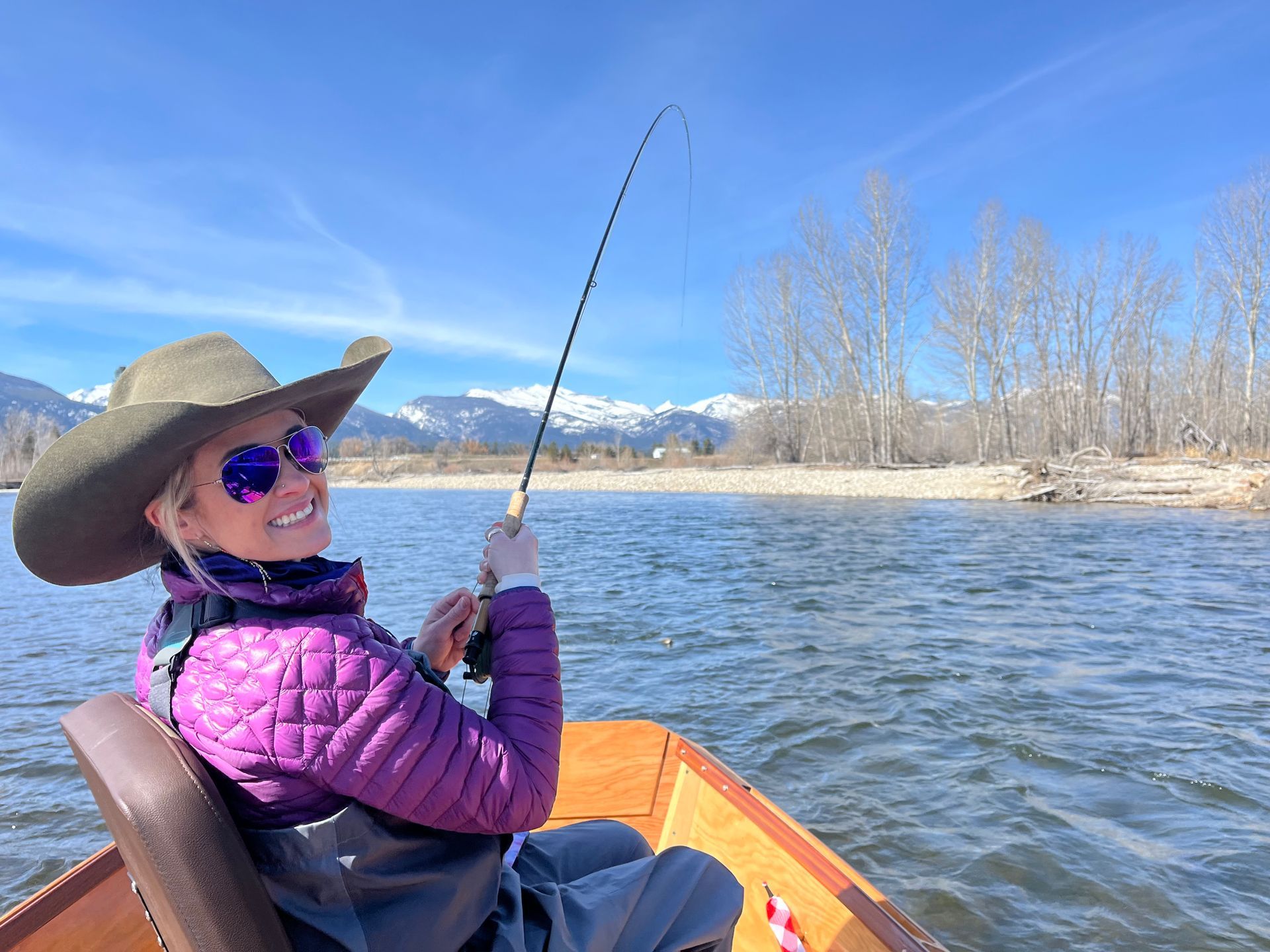 Woman in cowboy hat fishing from a boat on a river, mountains in background.