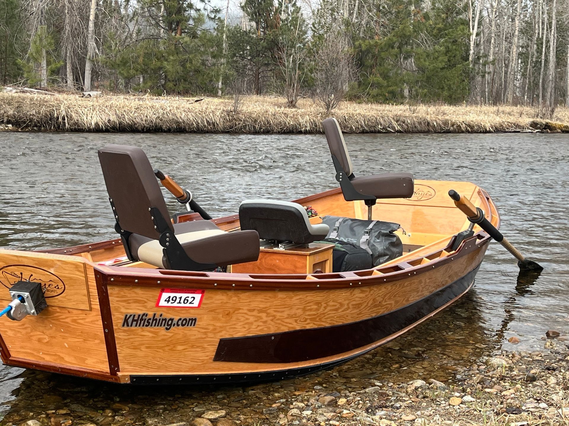 Wooden drift boat on a sandy riverbank, mountains and trees in the background.