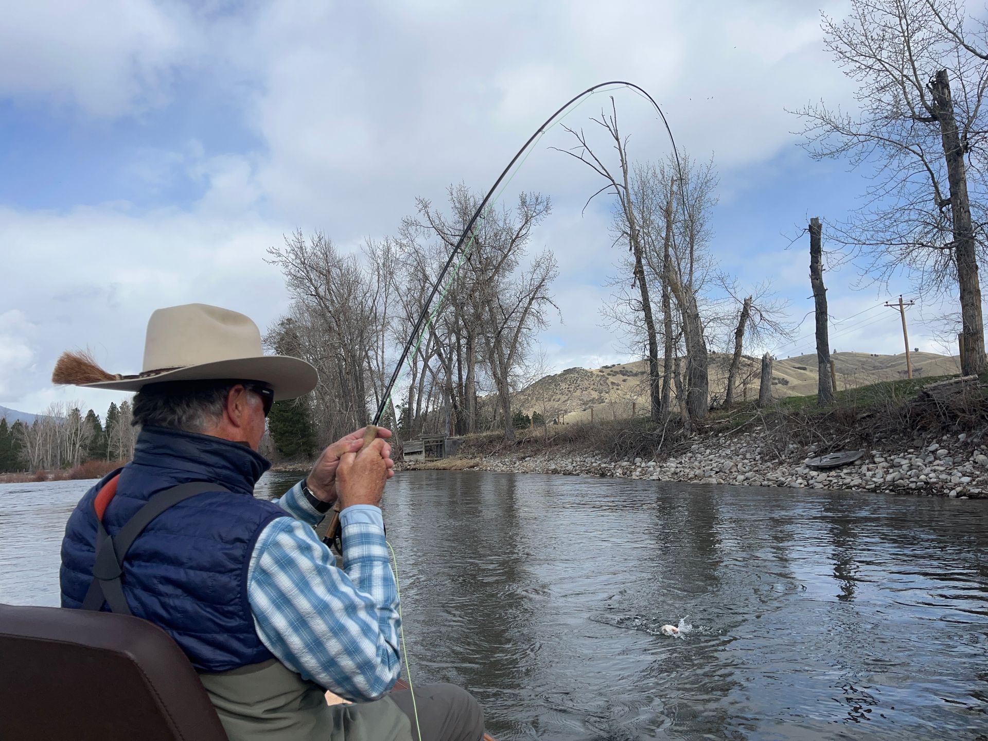 Angler fishing from a boat on a river, rod bent, wearing a cowboy hat and vest.