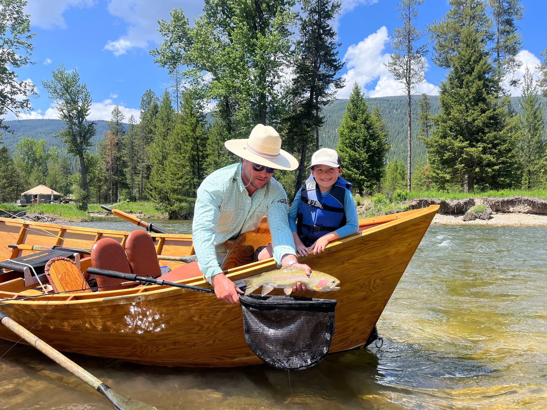 Man and child in a wooden boat hold a netted fish in a river, trees in background on a sunny day.