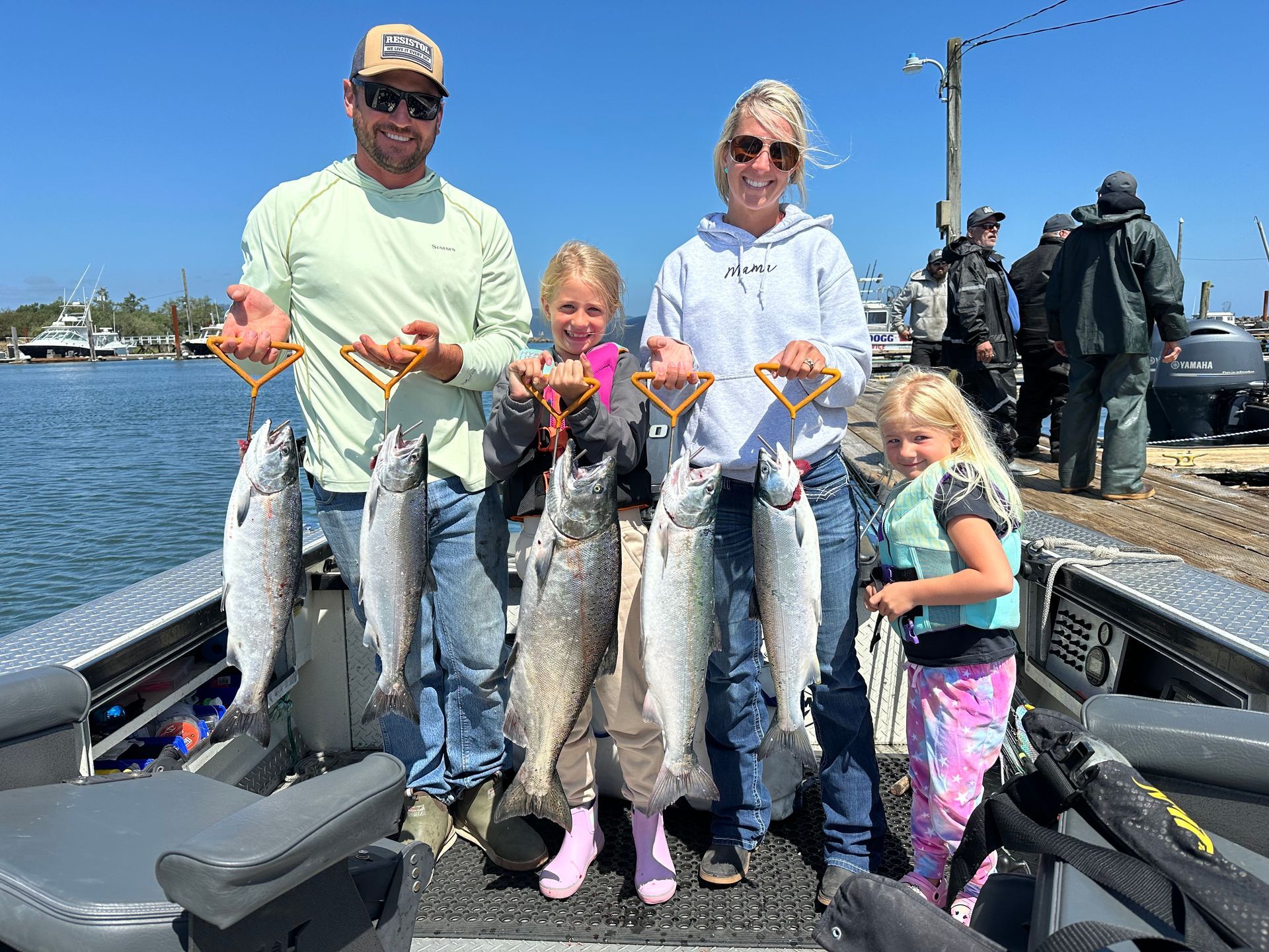Family on a boat, holding up several fish they caught. Bright sunny day by a dock.