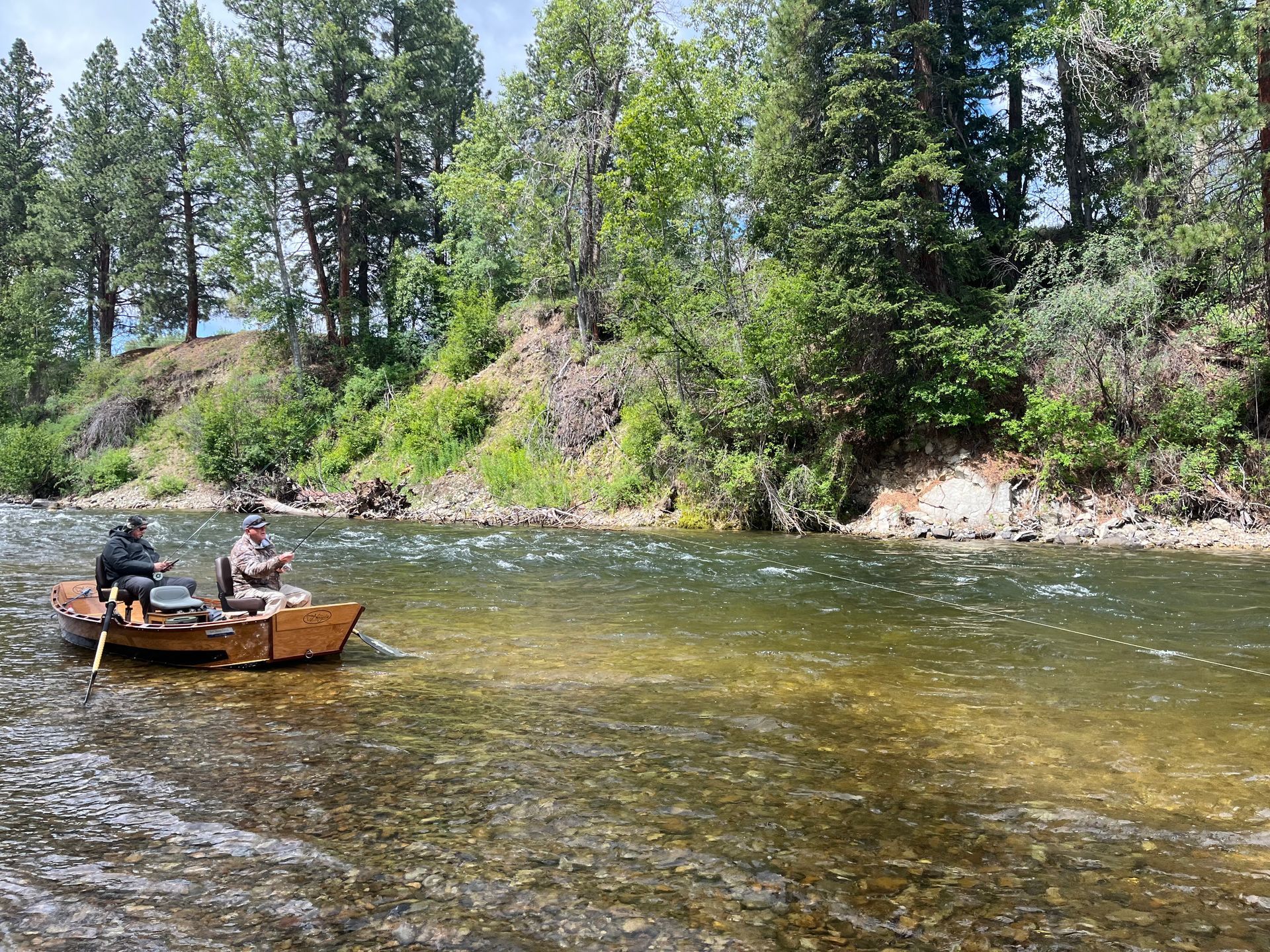 Two people fishing from a wooden boat in a river lined with trees.