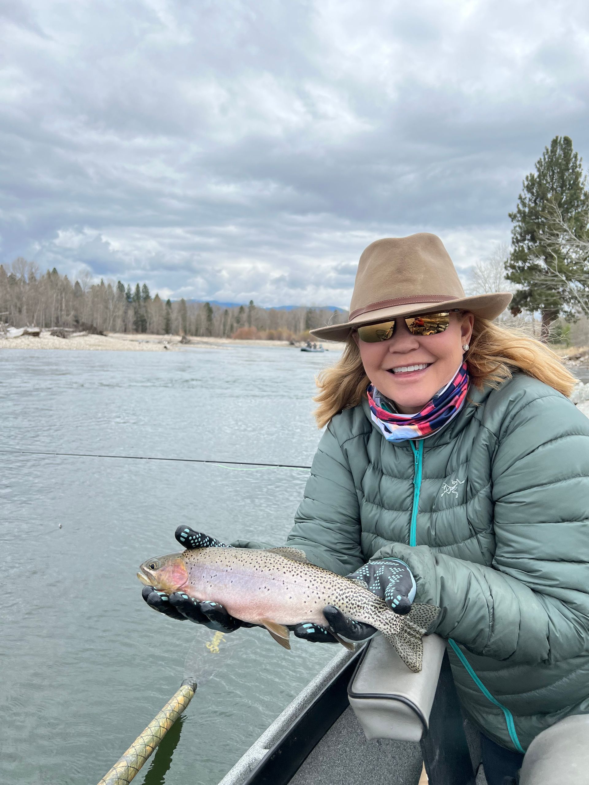 Woman holding a rainbow trout, smiling. Cloudy day, river in background.