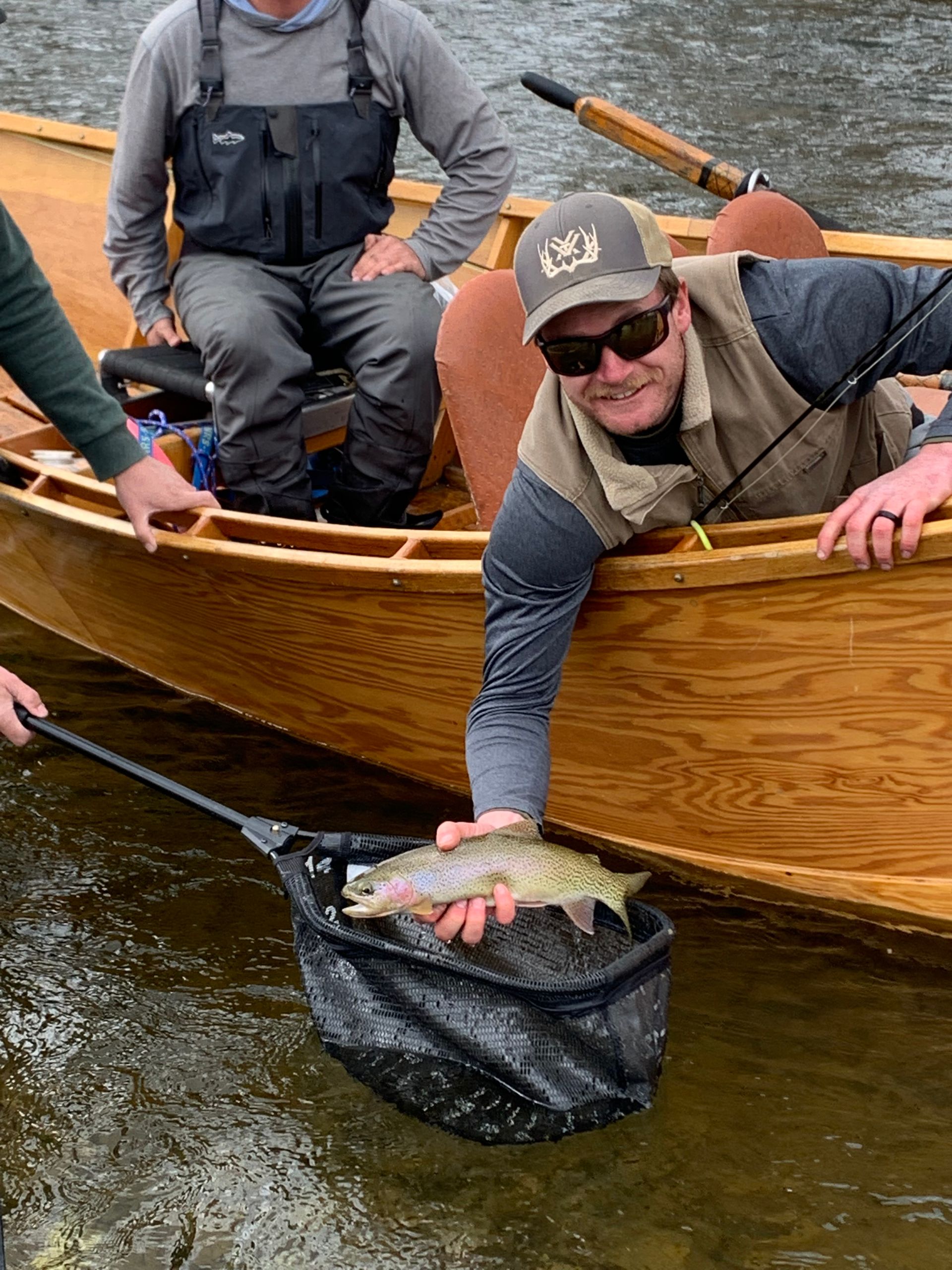 Man holding fish, smiling, from wooden boat on water. Another man nearby. Net in foreground.