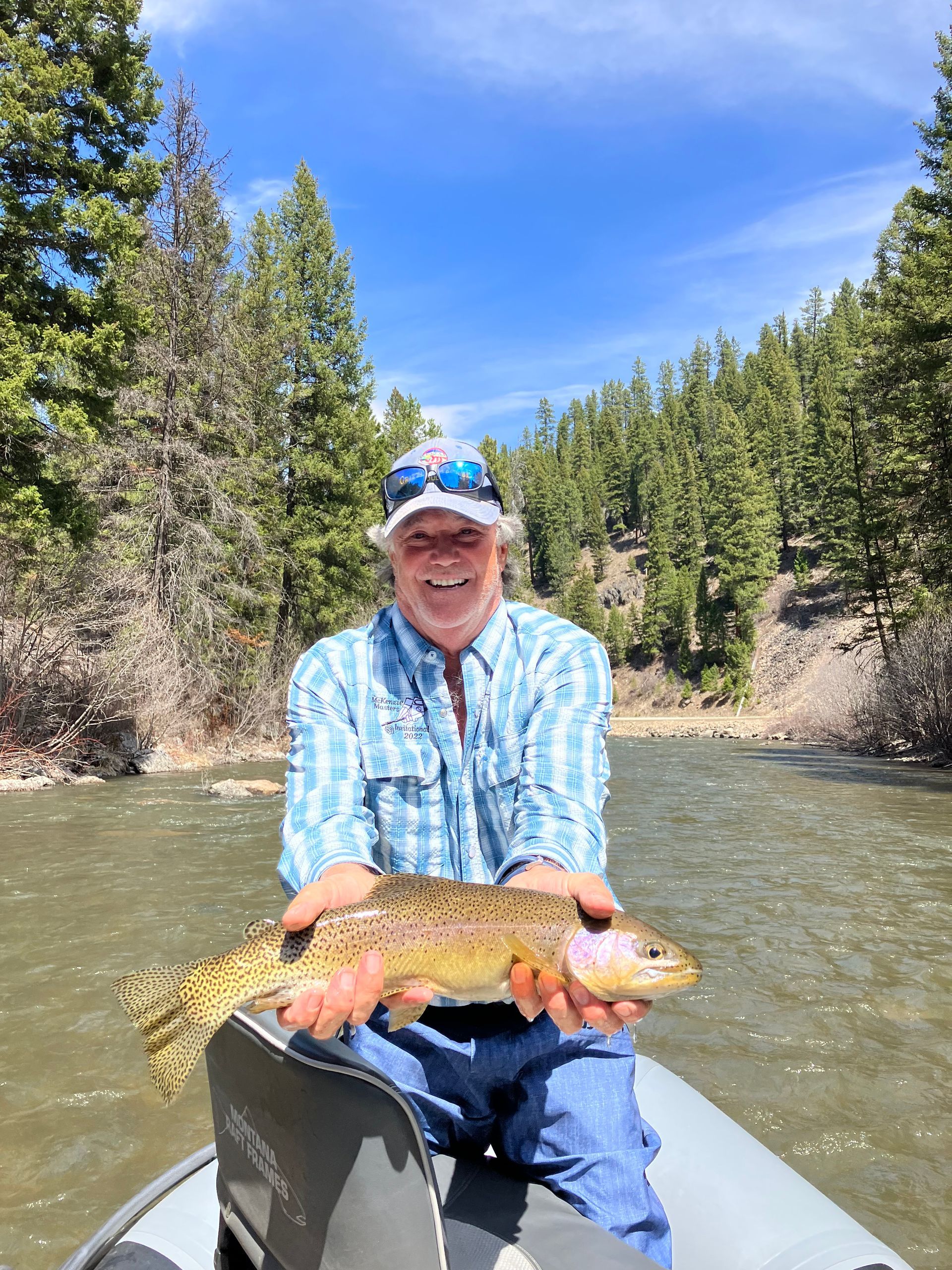 Man holding a large trout on a boat, smiling, with a river and trees in the background.