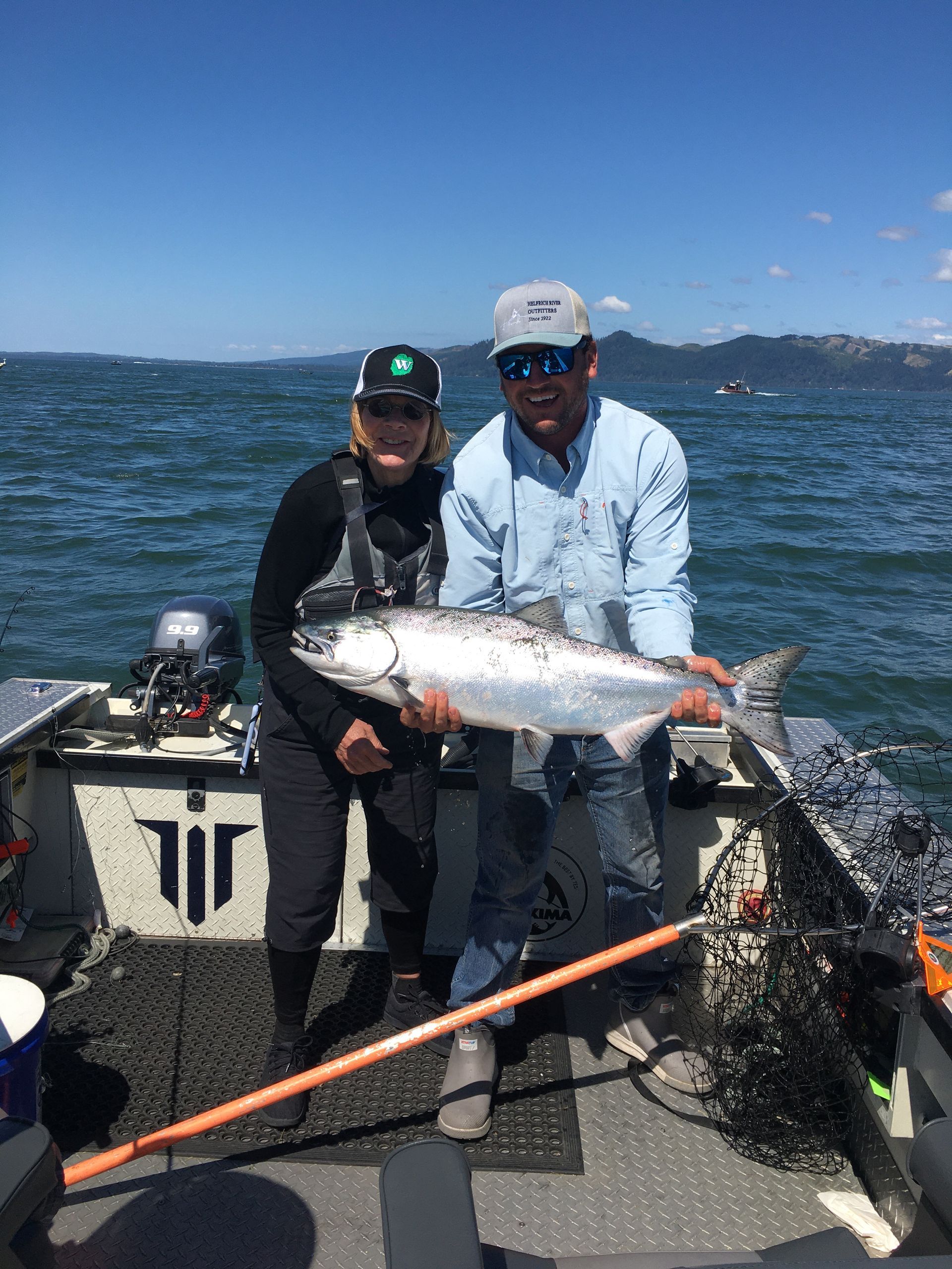 Two people on a boat holding a large silver fish. Blue water and sky. Sunny day.