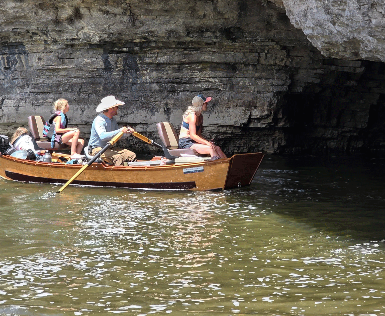 People in a wooden boat, rowing into a cave opening. Sunny day, water is choppy.