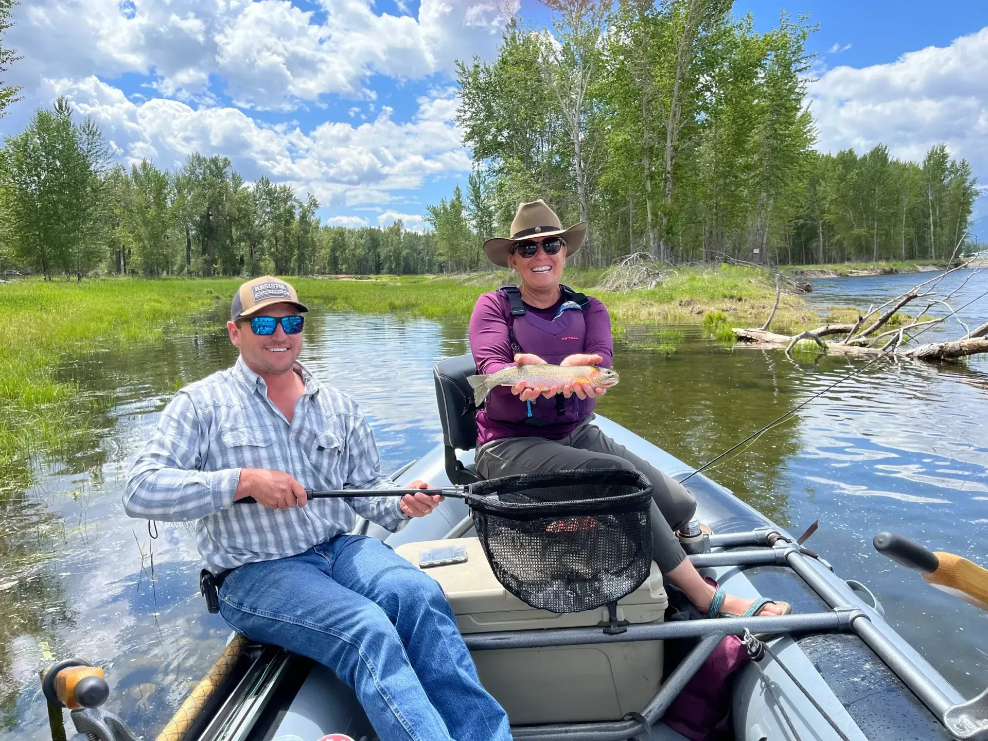 Two people fishing from a boat on a river, one holding a fish. Sunny day, green trees in background.
