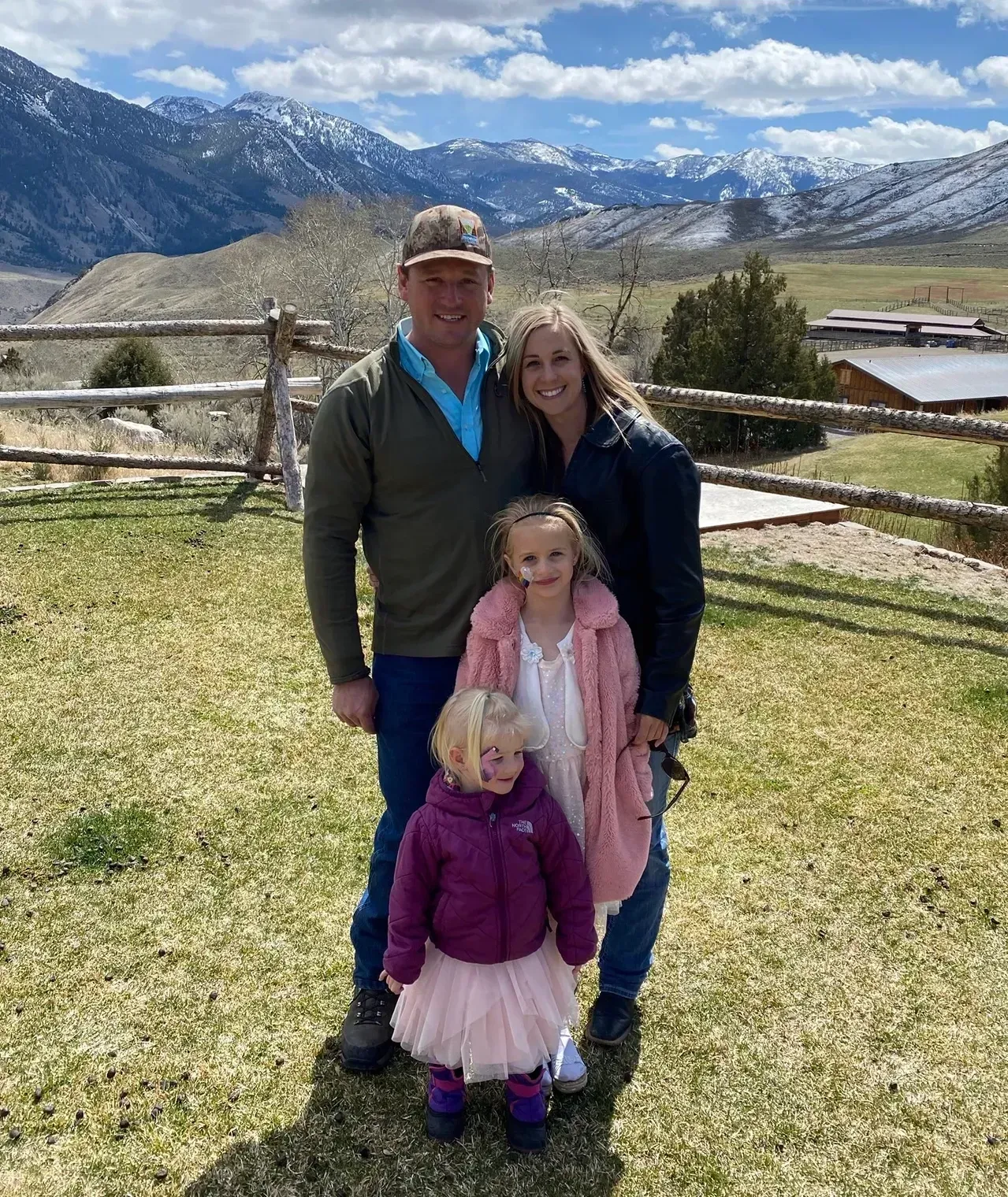 Family of four poses outdoors in front of mountains and a wooden fence.