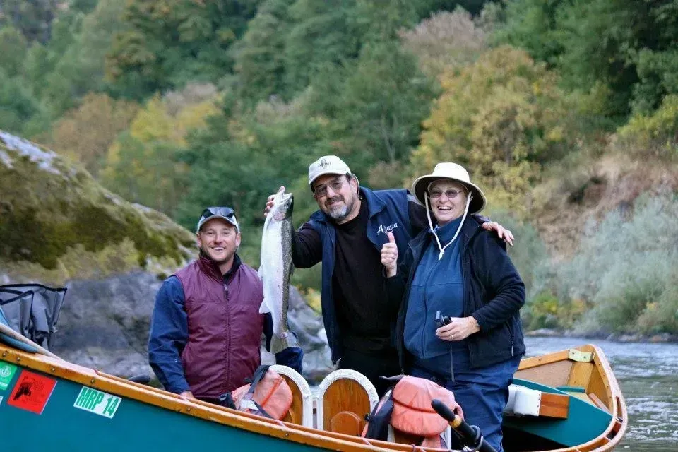 Three people in a boat holding a fish, near a river with autumn foliage.