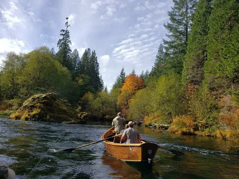 Two people rowing a wooden boat on a river surrounded by trees with fall colors.