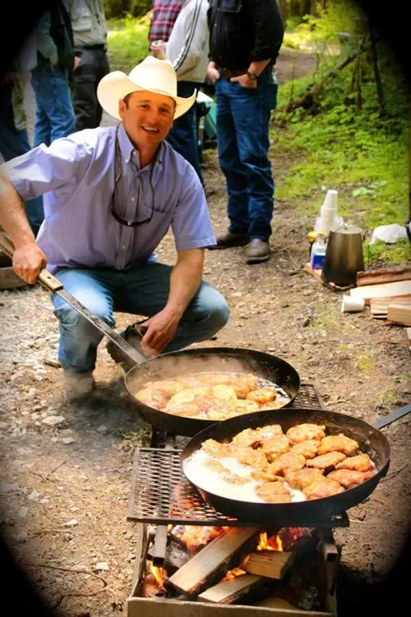Man in cowboy hat cooking food in two pans over a fire outdoors, smiling.