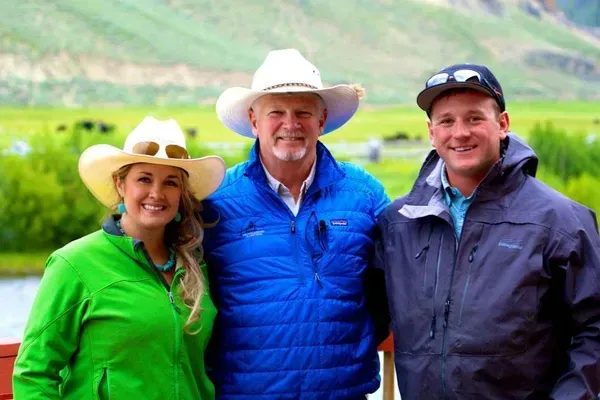 Three people pose outdoors, smiling. Two in cowboy hats, one in a baseball cap and jackets, with a mountainous backdrop.
