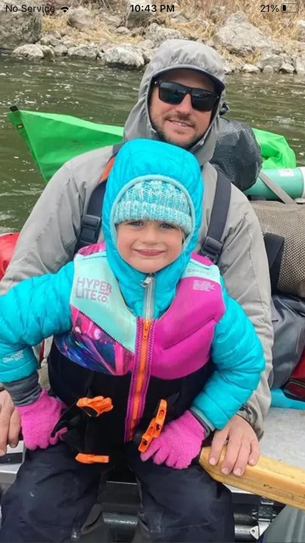 Man and young child in a raft on a river, both wearing warm outerwear and life vests.